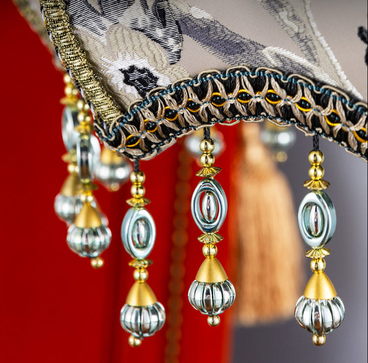 Close-up detail shot of an ornate traditional Chinese-style lamp shade with hanging bead tassels, embroidered fabric, and gold decorative trim against a red background. Chinese text appears on the image.