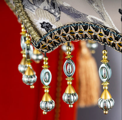 Close-up detail shot of an ornate traditional Chinese-style lamp shade with hanging bead tassels, embroidered fabric, and gold decorative trim against a red background. Chinese text appears on the image.
